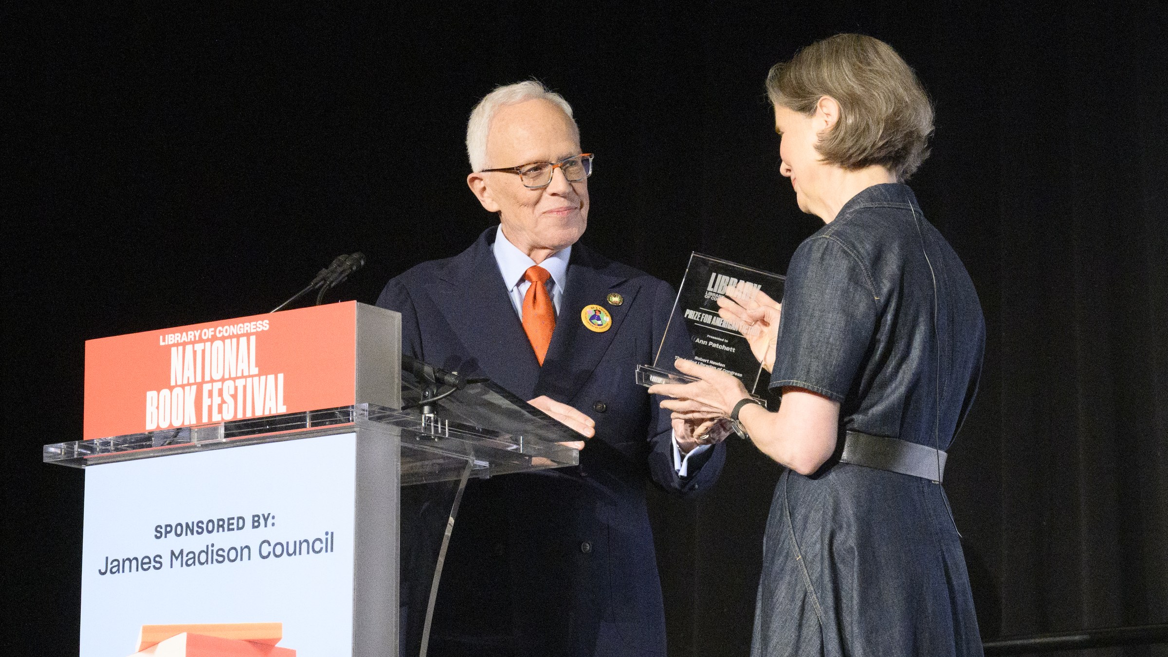 Robert R. Newlen, in a navy suit and orange tie, hands a glass award to Ann Patchett beside a National Book Festival lectern