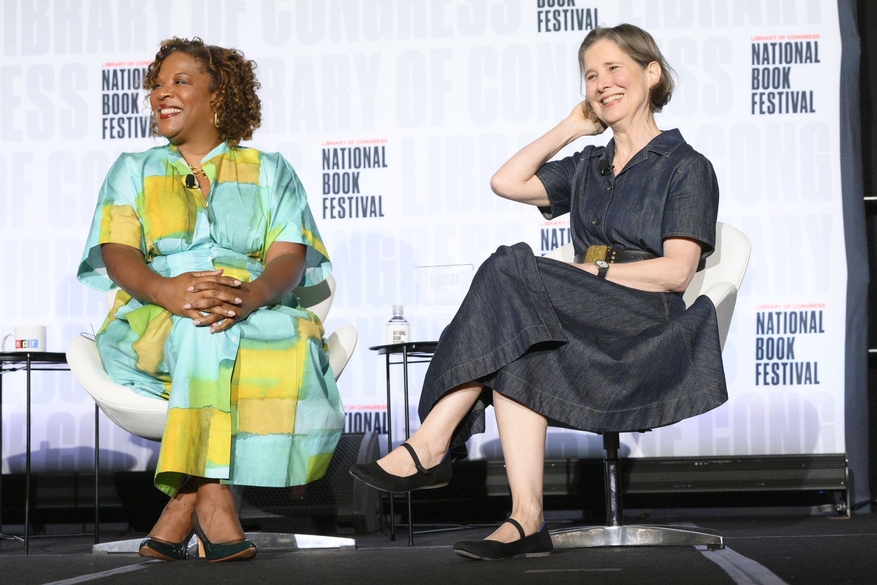 Tayari Jones in a green and yellow dress and Ann Patchett in a denim dress, both laughing in white chairs on a festival stage