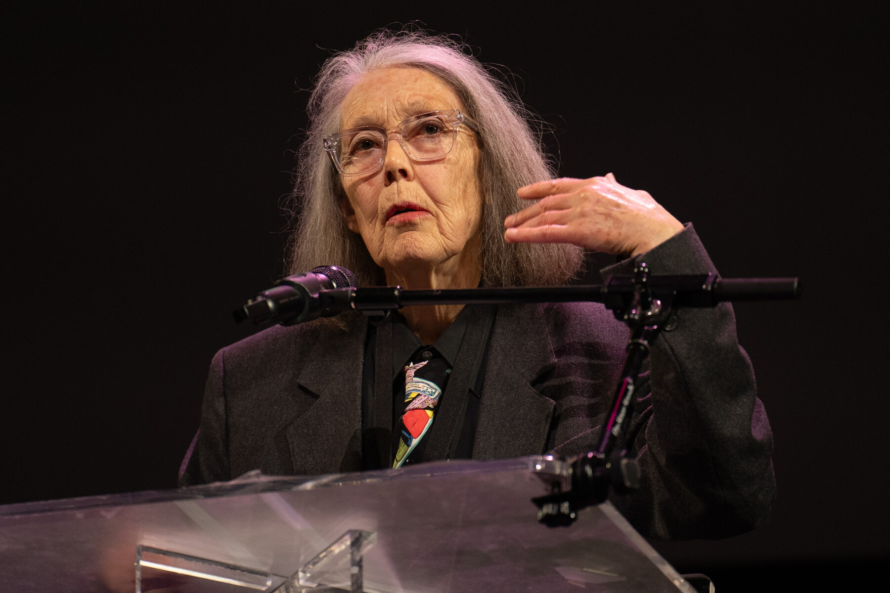 Anne Carson reading at a clear lectern on a dark stage