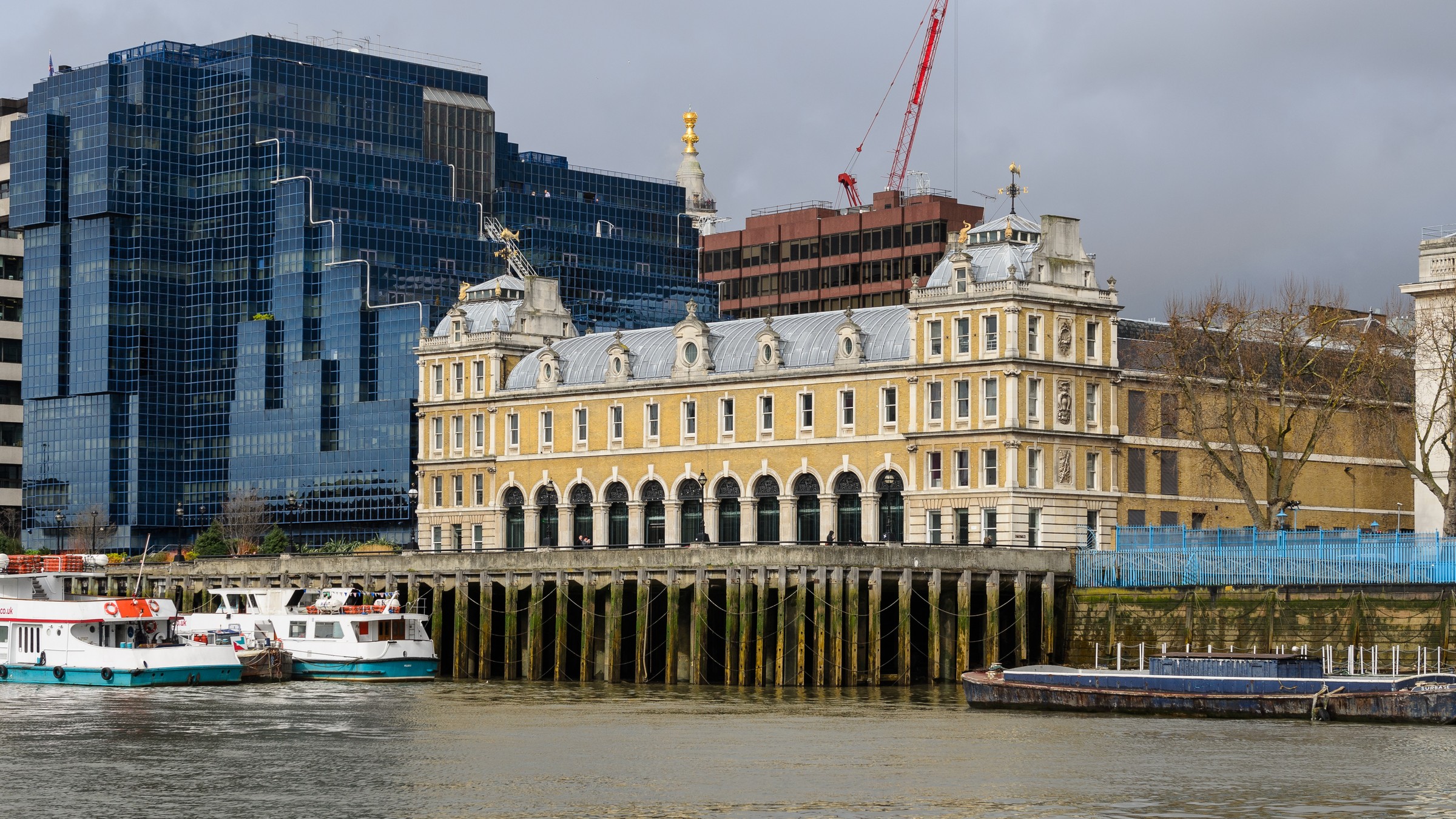 Old Billingsgate, a grand Victorian market hall in pale yellow brick with arched windows, seen across the Thames under a grey sky