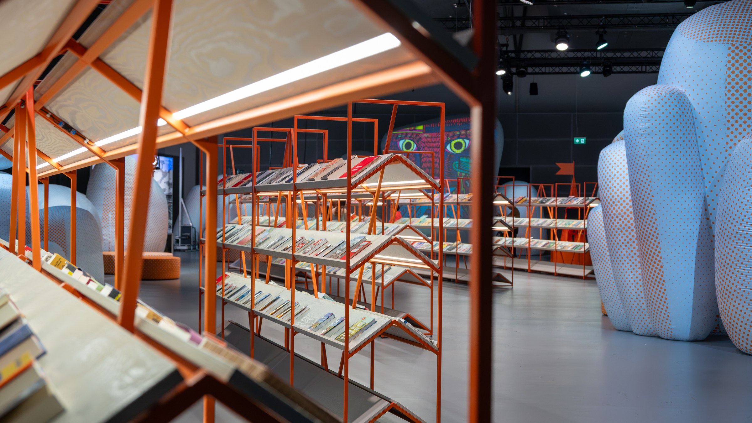 Orange metal bookshelves shaped like little house roofs, lined with Czech books, beside tall white inflatable forms in a dark hall