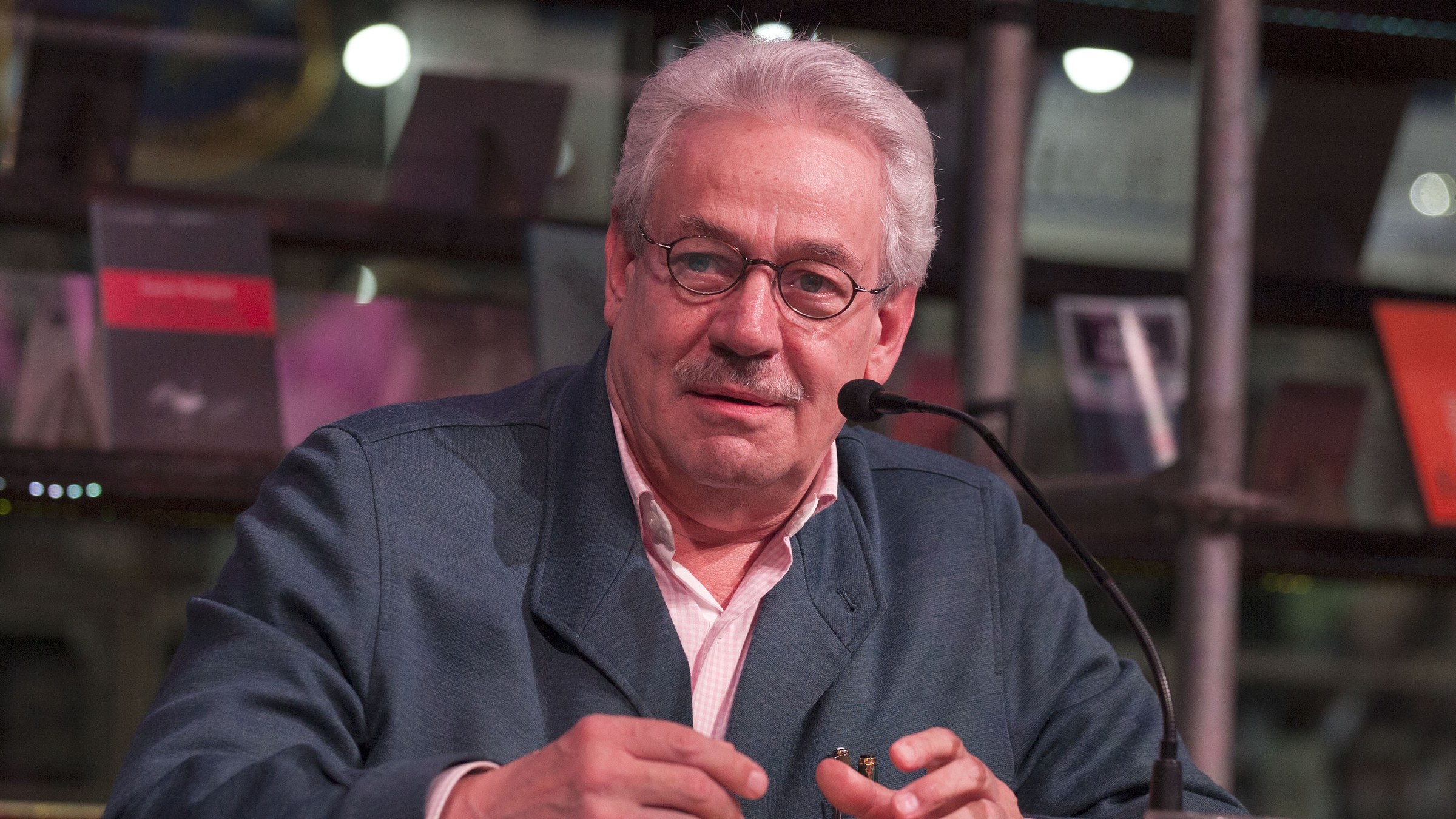 Gonzalo Celorio, white-haired, in glasses and a blue jacket, smiling at a microphone in front of bookshelves