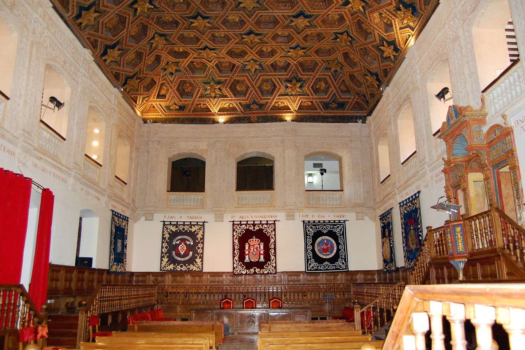 A long hall with white walls, red-and-gold banners and a richly carved, gilded wooden ceiling above rows of wooden benches