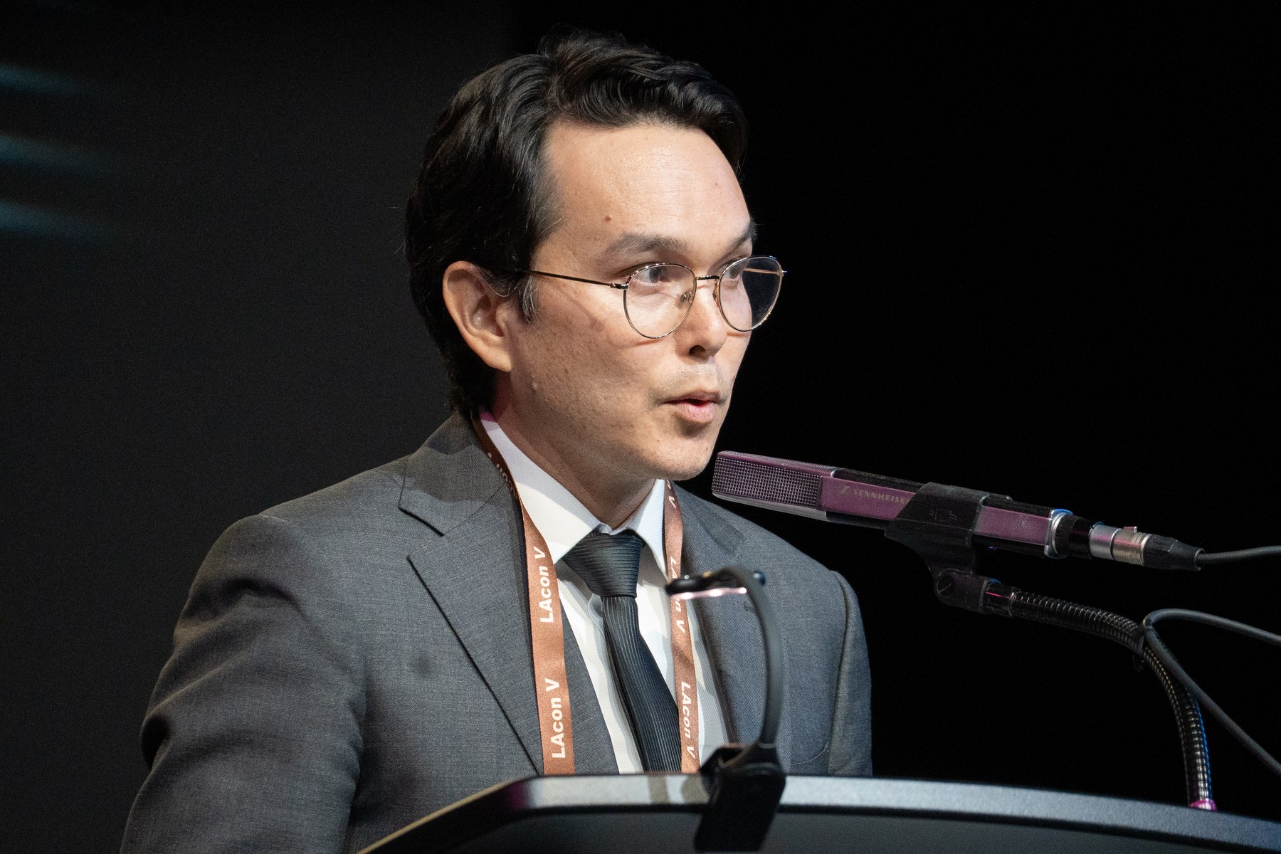 Thomas Ha, in glasses and a gray suit, speaking into a microphone at a lectern on a dark stage