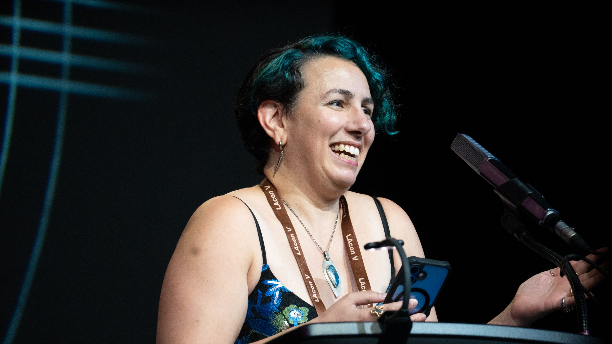 Amal El-Mohtar, with short teal-tinted hair, laughing at a lectern with a phone in her hand on a dark stage