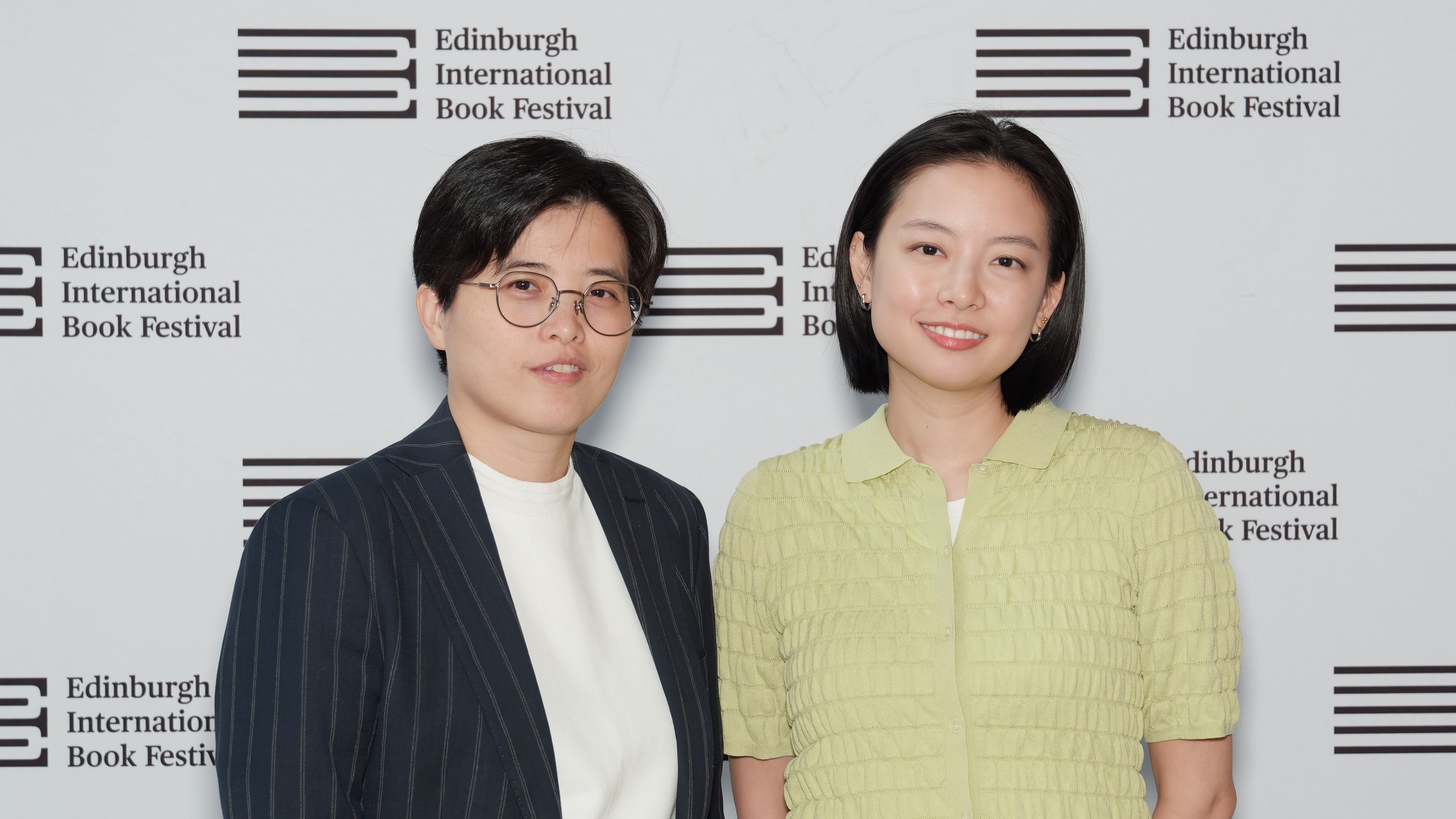 Yáng Shuāng-zǐ, in glasses and a pinstriped jacket, stands beside Lin King, in a green blouse, in front of an Edinburgh International Book Festival backdrop
