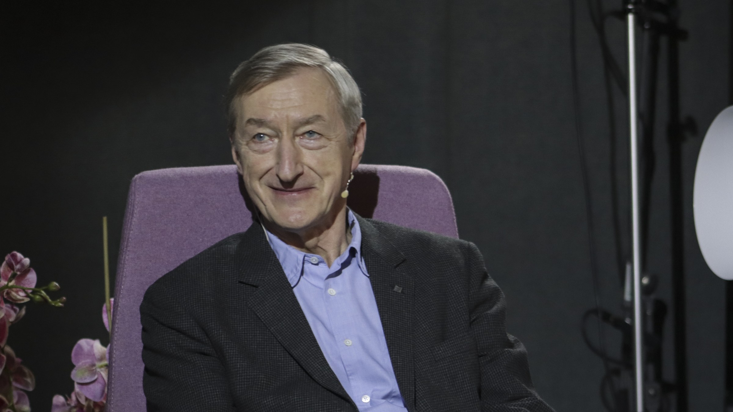 Julian Barnes, in a dark jacket and blue shirt, smiling in a lilac armchair on a dimly lit stage