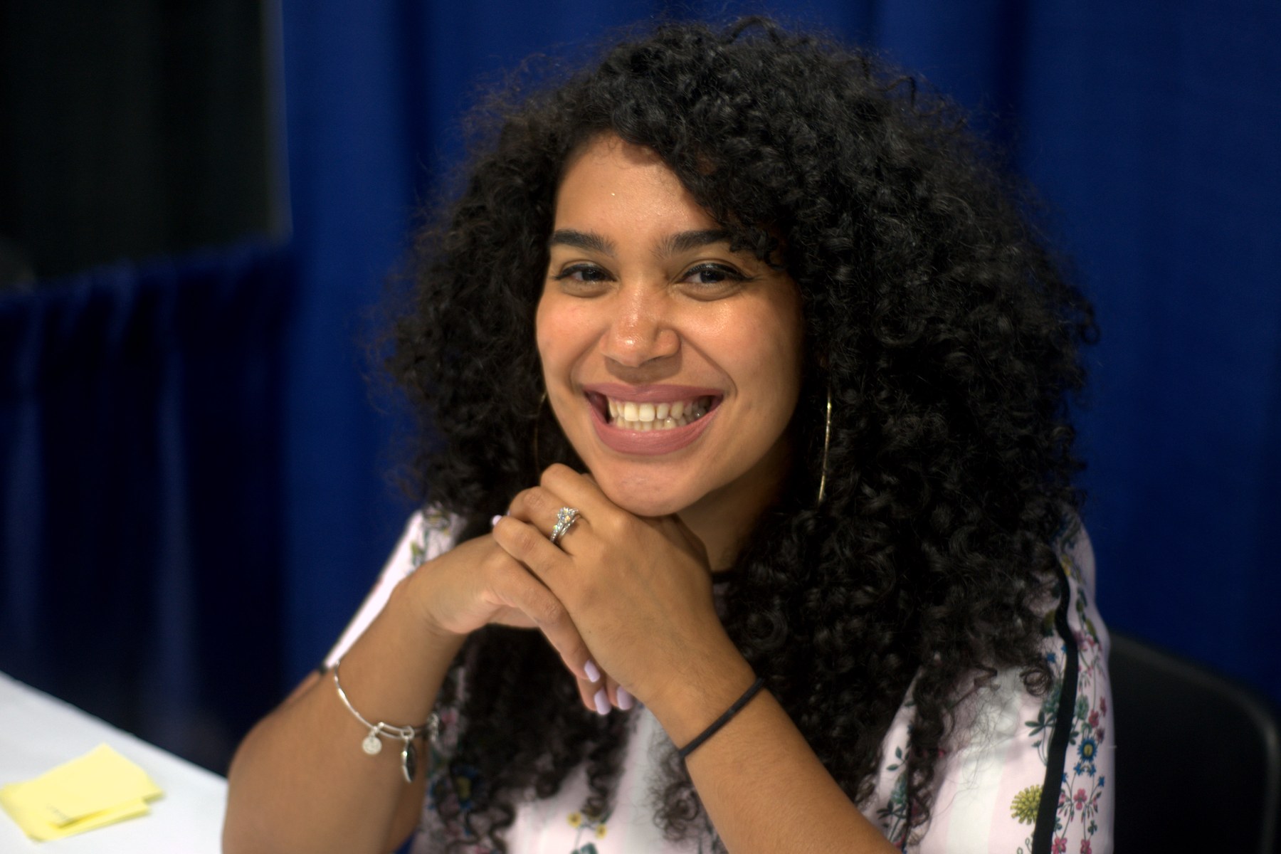 Elizabeth Acevedo, with long dark curly hair, smiling with her chin resting on her hands in front of a blue curtain