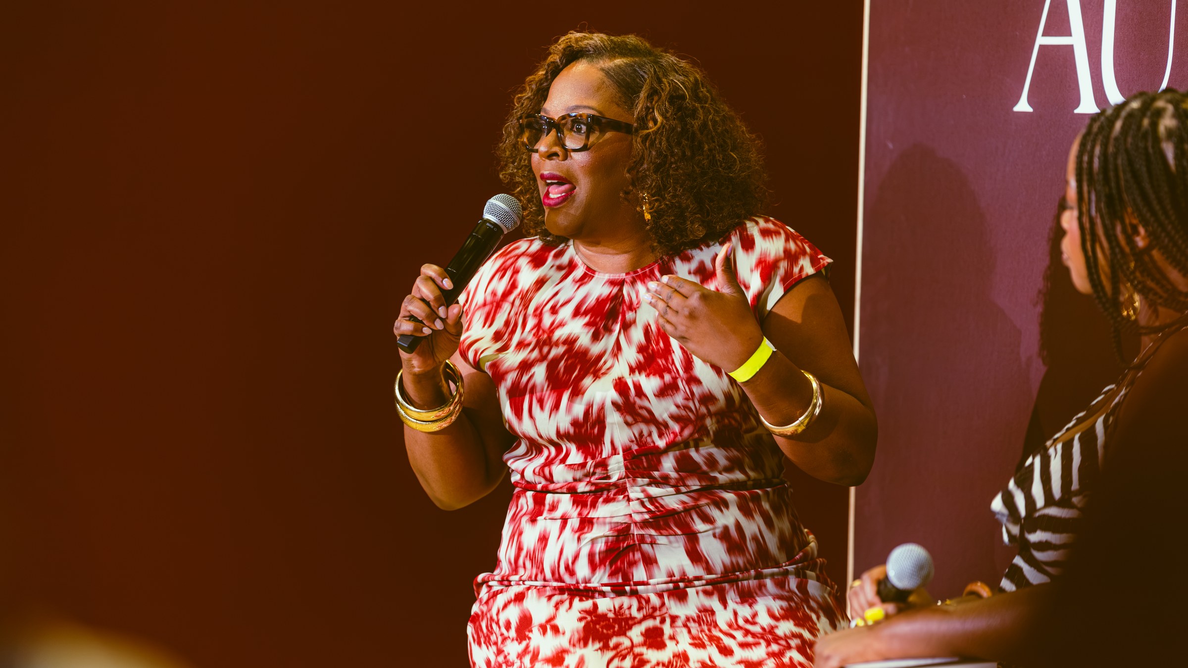 Tayari Jones, in tortoiseshell glasses and a red and white dress, speaking into a microphone on a dark red stage