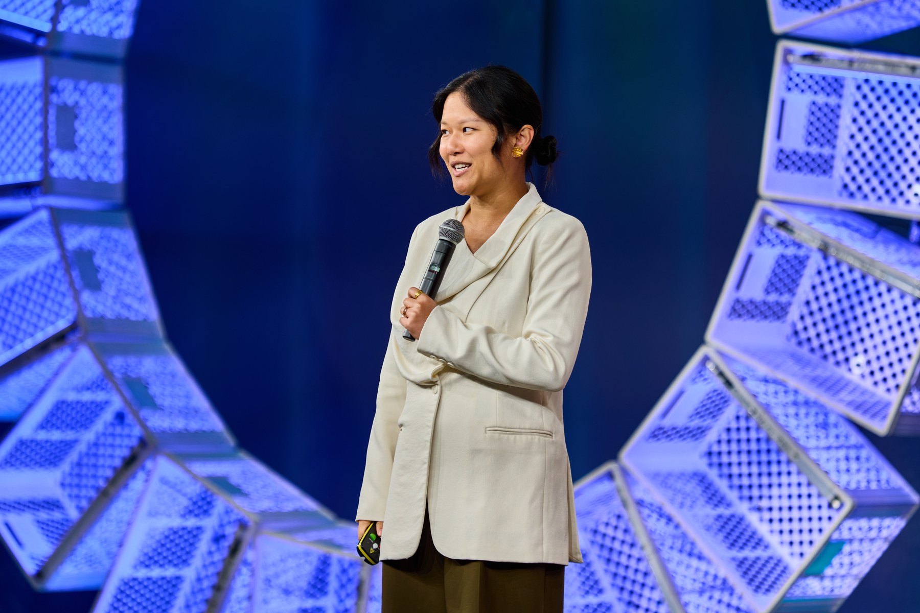 Karen Hao in a cream jacket, holding a microphone and smiling, on a stage ringed with glowing blue panels
