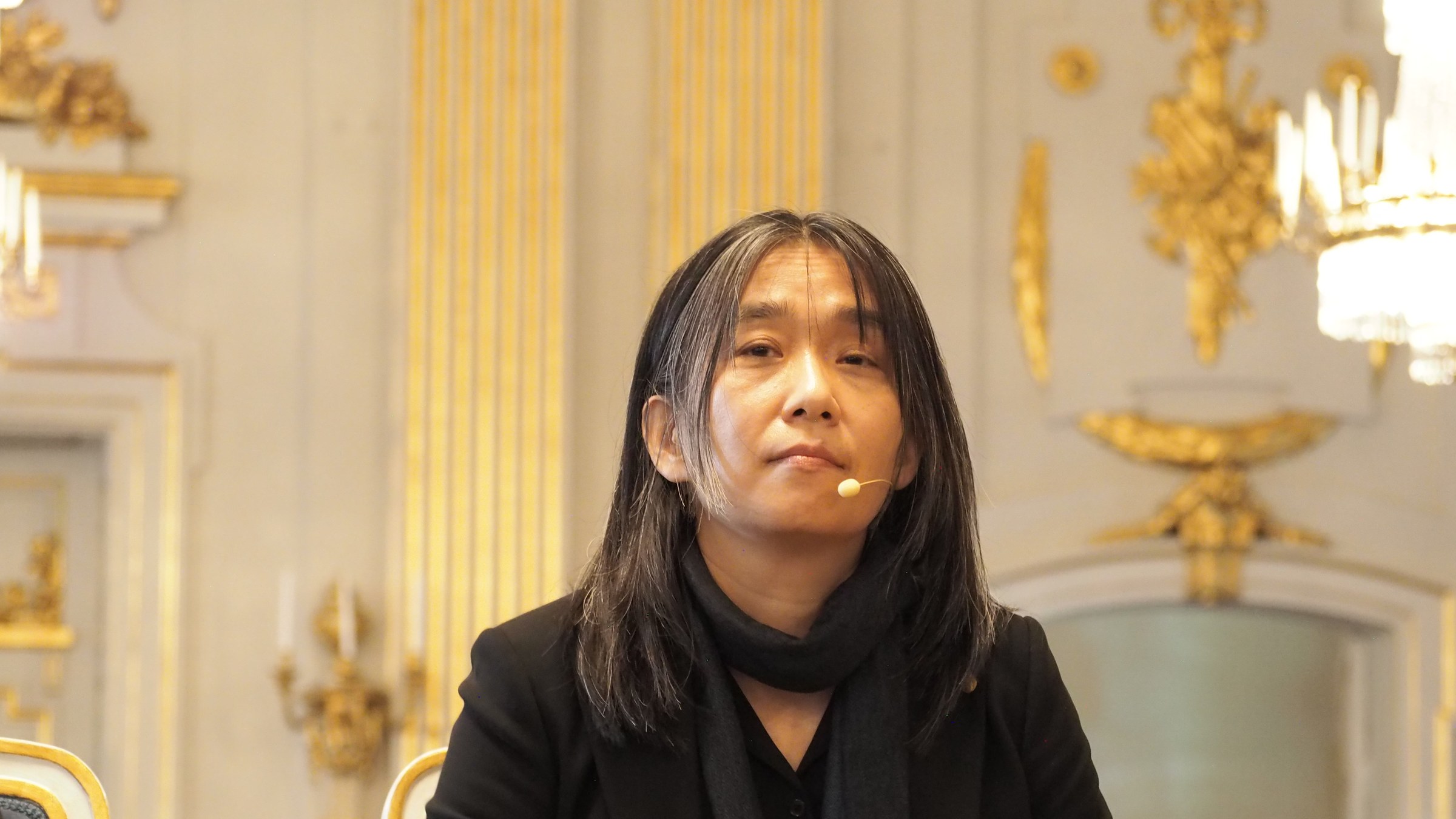 Han Kang, with long dark hair and a black scarf, listening at a press conference in a gilded hall