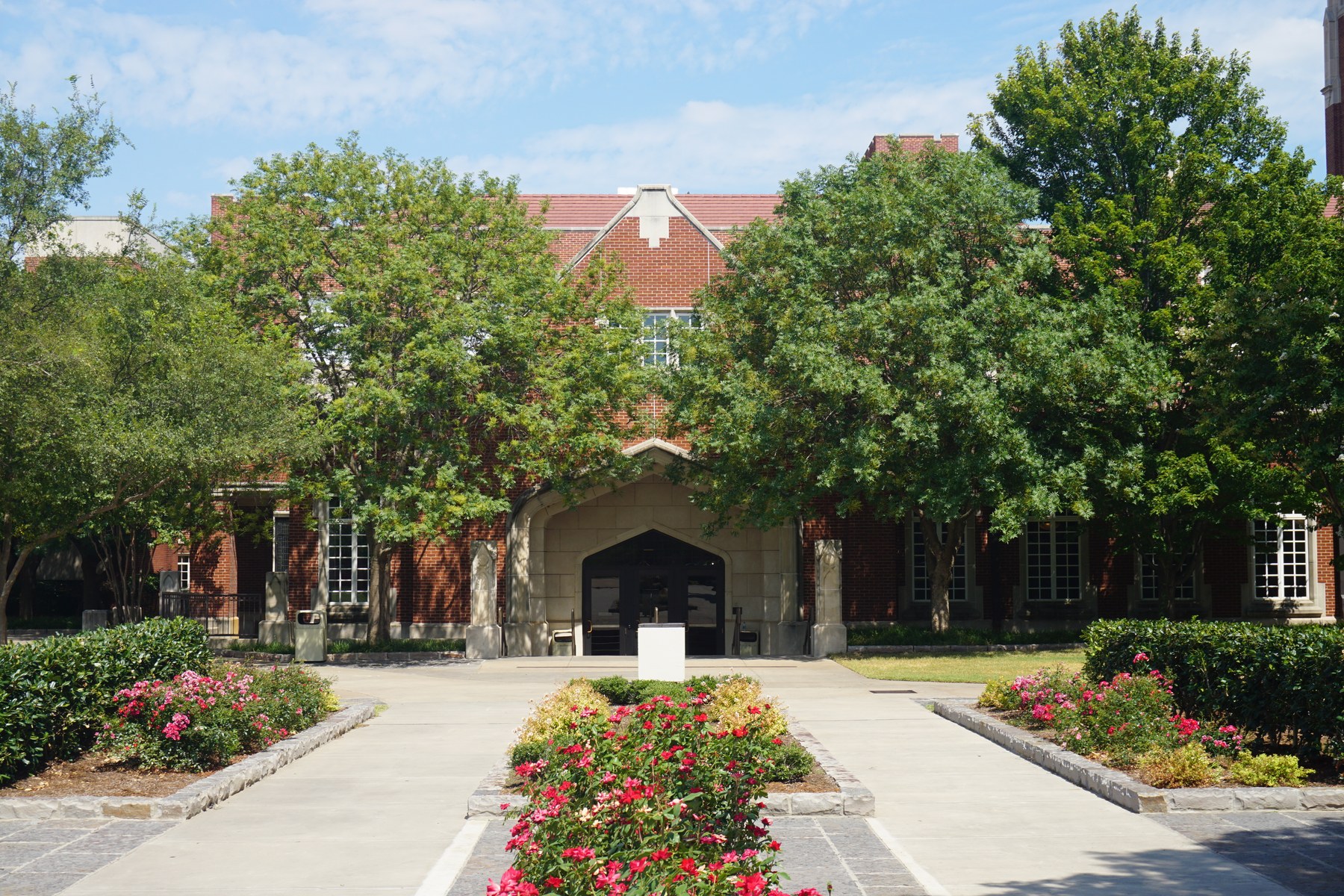 A red brick Gothic-style building with a tall square tower, seen across a flower-lined walkway and trees on a sunny day