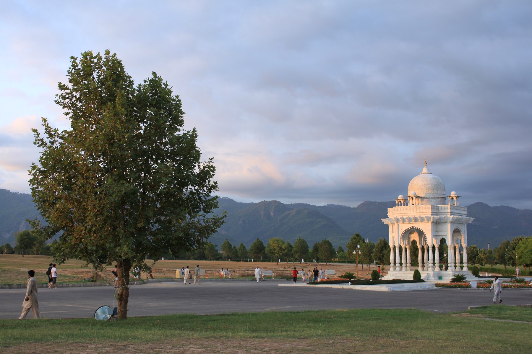 A wide lawn with a single tree, a white domed pavilion and green hills under an evening sky in an Islamabad park