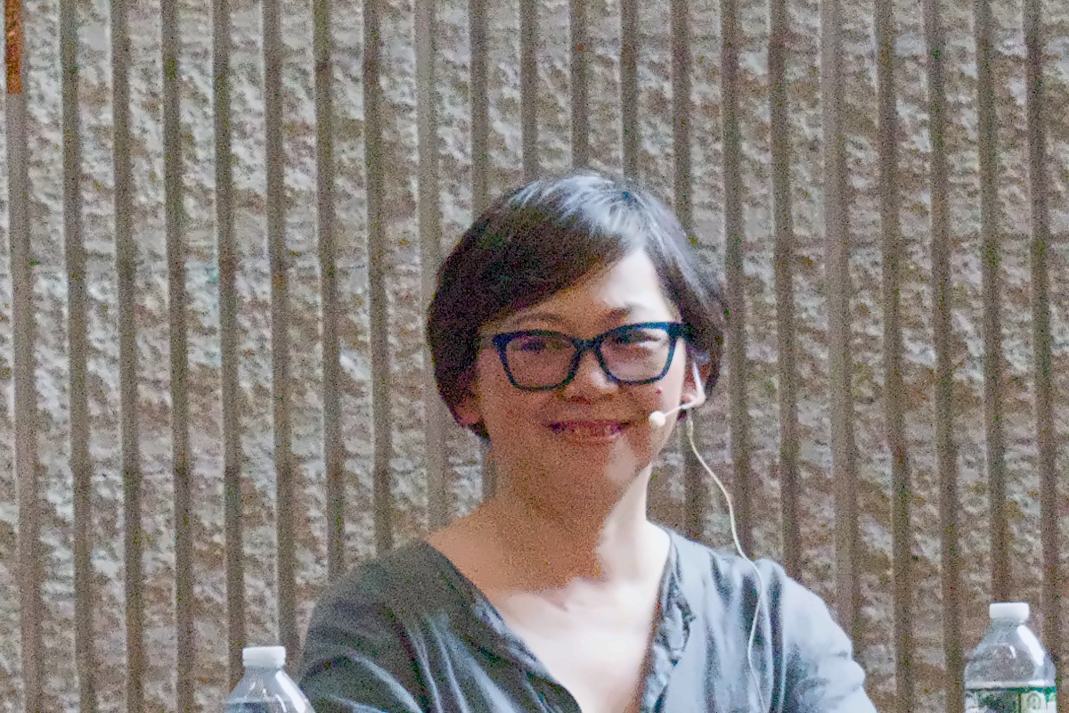 Yiyun Li smiling, in dark glasses, seated at a table on a festival stage