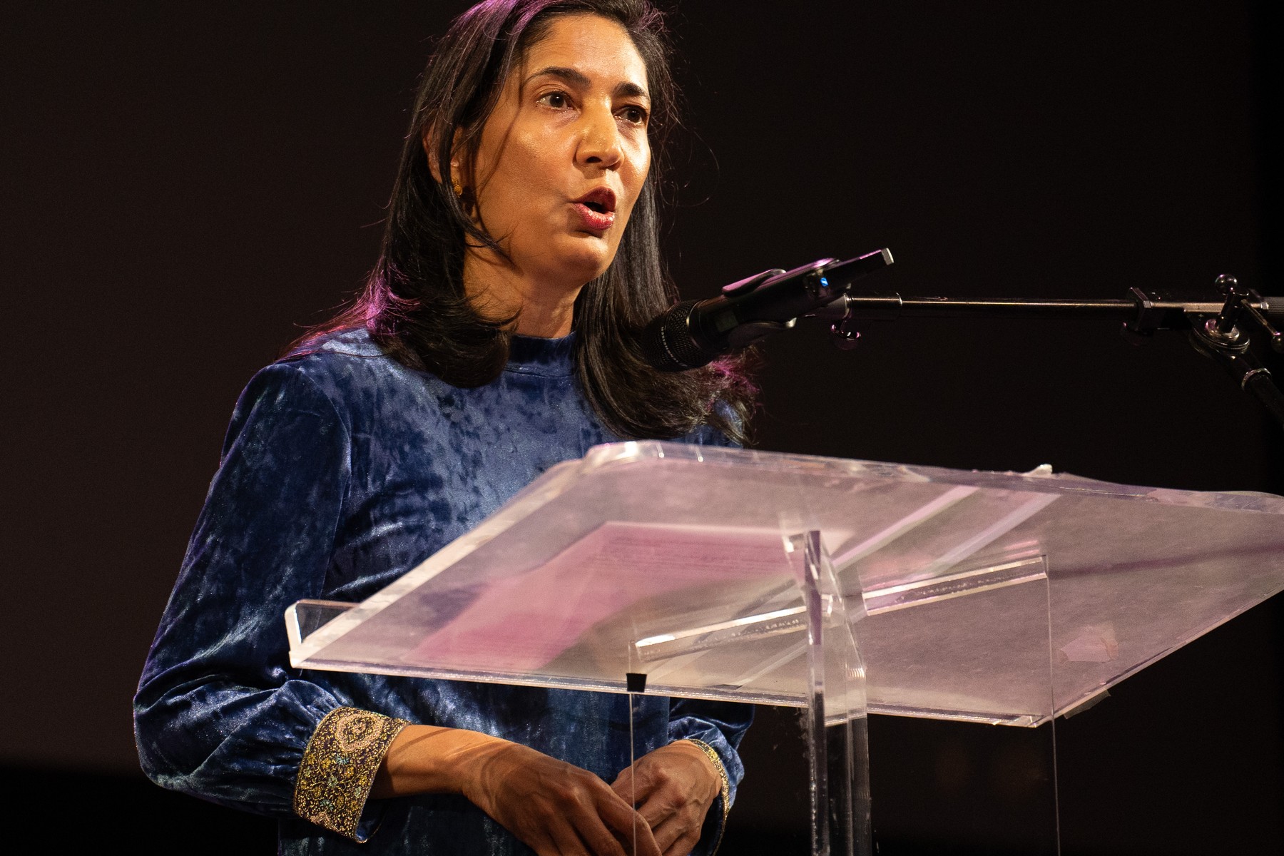 Kiran Desai, in a blue velvet dress, speaking at a clear lectern on a darkened stage