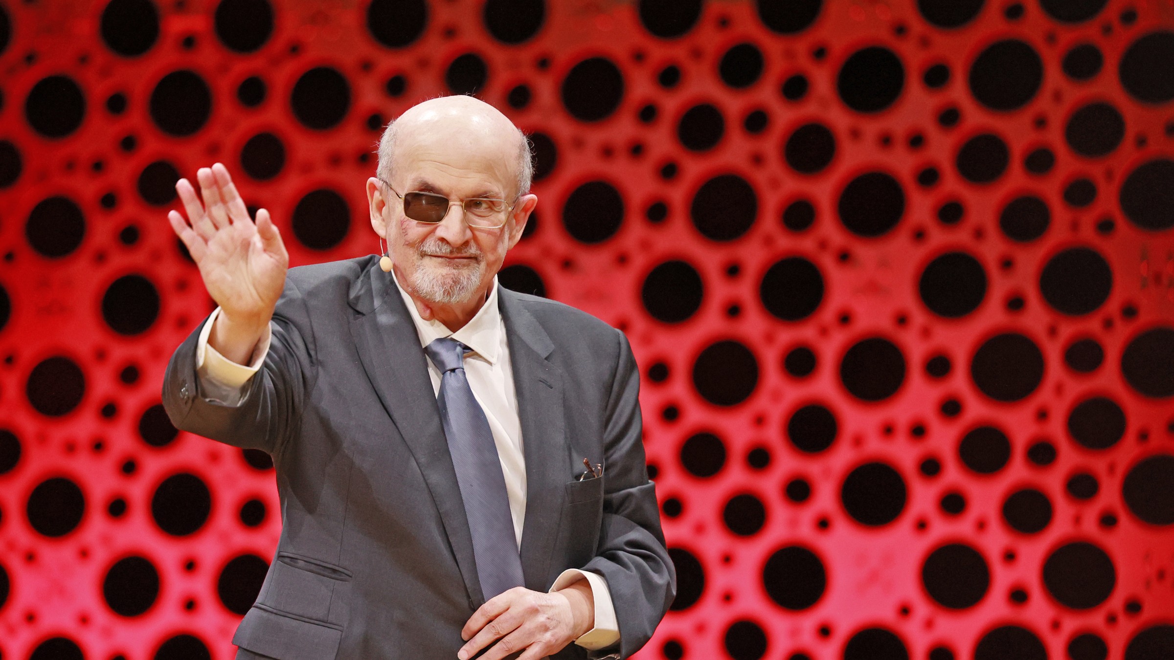Salman Rushdie in a grey suit and tinted glasses, waving to an audience in front of a red and black patterned backdrop