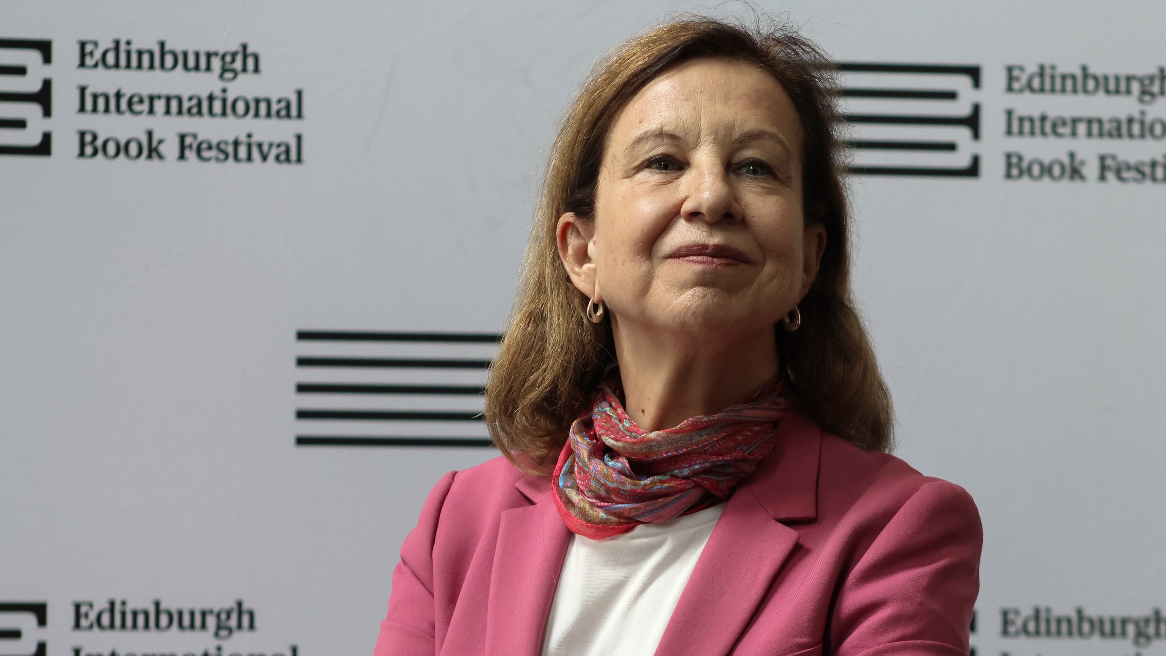 Lyse Doucet, in a pink jacket and patterned scarf, smiling in front of an Edinburgh International Book Festival backdrop