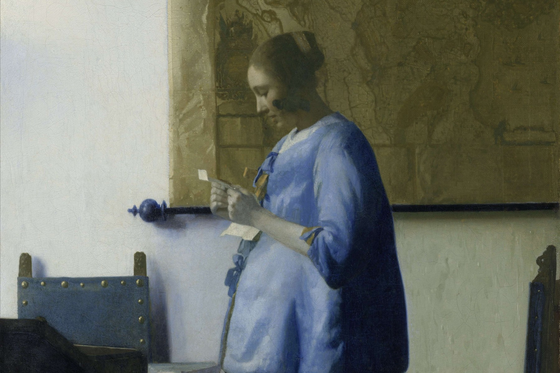 A Vermeer painting of a young woman in a blue jacket standing by a map on the wall, reading a letter held in both hands